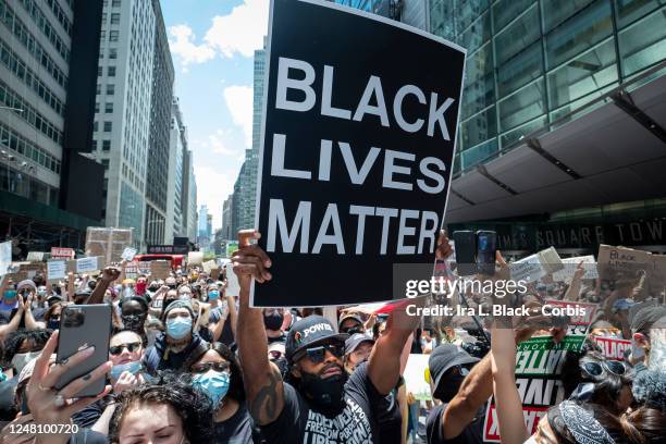 An African American protester wearing a mask around his neck and a hat that has Black Power in Times Square holds up a large sign that reads, "Black...