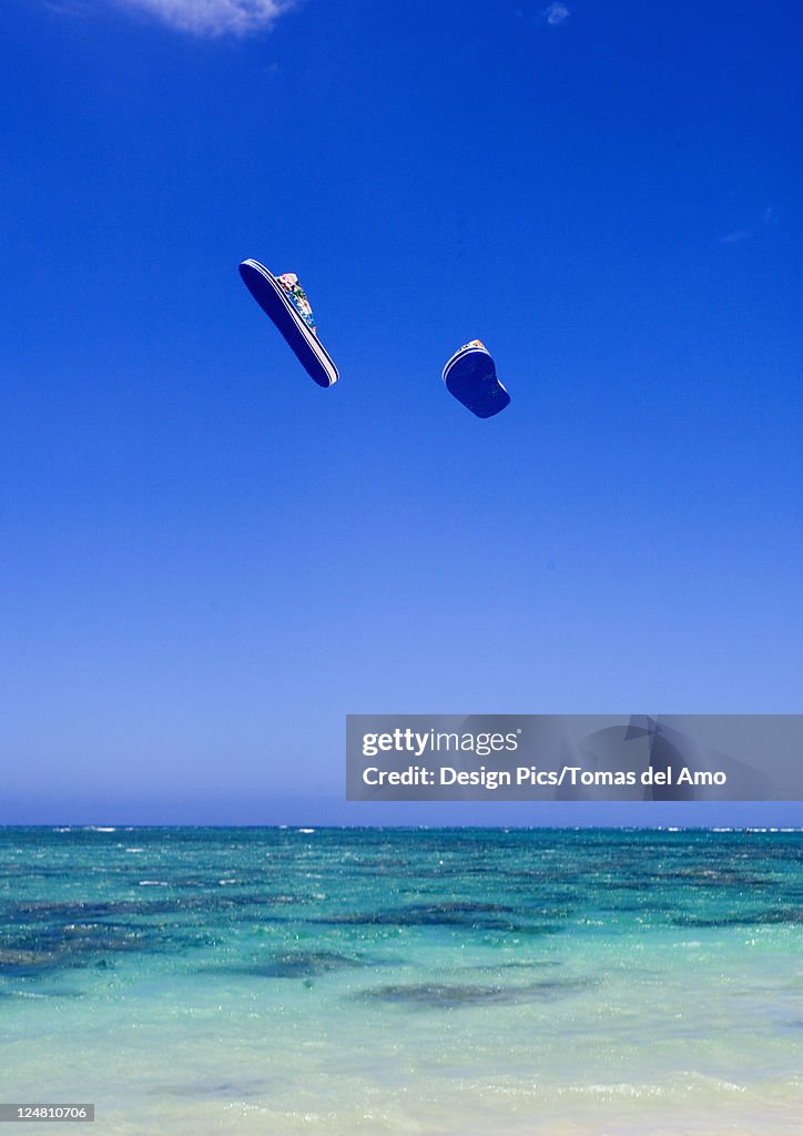 A pair of flipflops flying through the air above turquoise ocean.