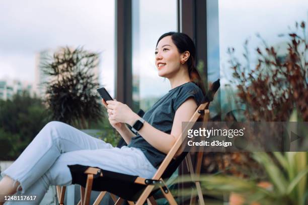 cheerful young asian woman using smartphone while relaxing on deck chair in the backyard, surrounded by beautiful houseplants - backyard vacations stock pictures, royalty-free photos & images