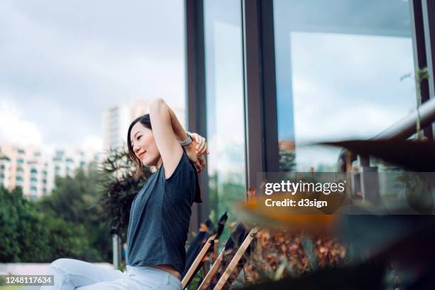 young asian woman stretching hands while relaxing on deck chair in the backyard, surrounded by beautiful houseplants - backyard deck stockfoto's en -beelden