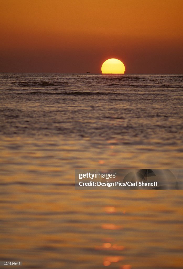 Sunball sinking into the ocean at sunset, red sky, color reflecting on water.