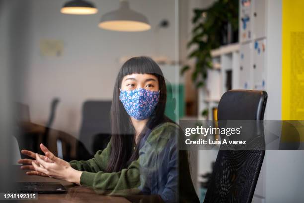 businesswoman discussing work with colleague through glass shield on desk - cloth face mask foto e immagini stock