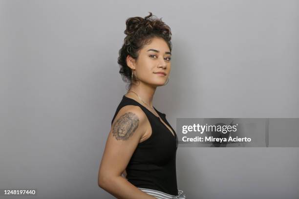 Profile of LatinX millennial woman with dark curly hair, wearing casual, summer clothing, with a serious expression, stands in front of a grey backdrop.