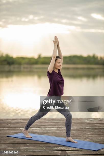 woman practicing yoga, standing in warrior one exercise, virabhadrasana i pose on lake pier - sun salutation stock pictures, royalty-free photos & images