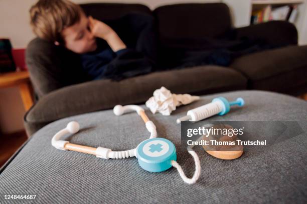 Symbolic photo on the subject of sick children: A five-year-old boy lies at home on a couch behind a stethoscope, a syringe and a handkerchief on...