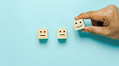 Top view of  Hand holding of Happy icon on cube blocks about rating feedback of survey from customer. Business annual survey concept. Many sad or happily on wooden on blue paper.