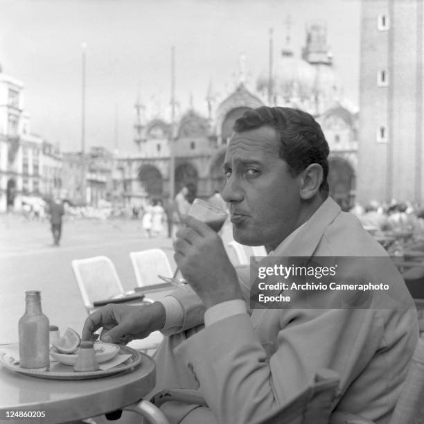 Italian actor Alberto Sordi, wearing a suit, portrayed while sipping a glass of tomato juice sitting on a terrace in St. Mark Square, salt, pepper...