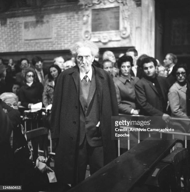 American poet Ezra Pound, wearing a suit, a tie and a coat, portrayed while standing next to Olga Rudge, the crowd behind him, Venice 1969.