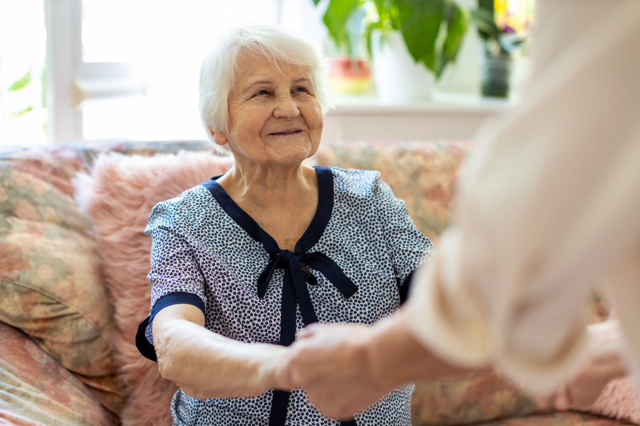 Female home carer supporting old woman to stand up from the sofa at care home Female home carer supporting old woman to stand up from the sofa at care home