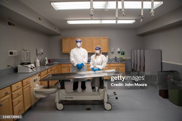 Funeral director Alexandra Burke and intern Vincent Belfiore, both wearing personal protective equipment stand at a mortuary table in the preparation...