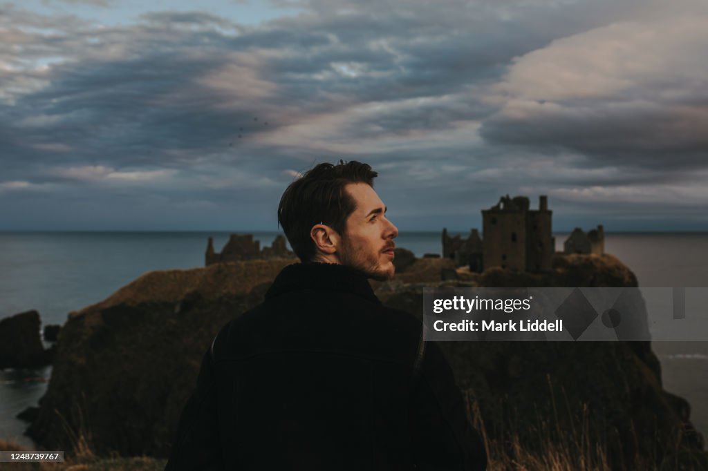 Young man standing in front of Dunnottar Castle at sunset, Scotland