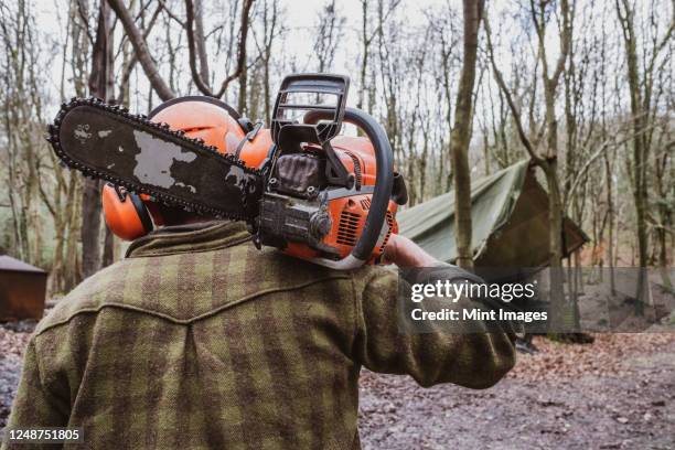 man wearing safety gear walking through forest carrying chainsaw on his shoulder. - sierra de cadena fotografías e imágenes de stock