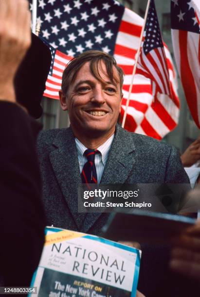 View of American author and political commentator William F Buckley Jr as he smiles, framed by several American flags, during an unspecified event,...