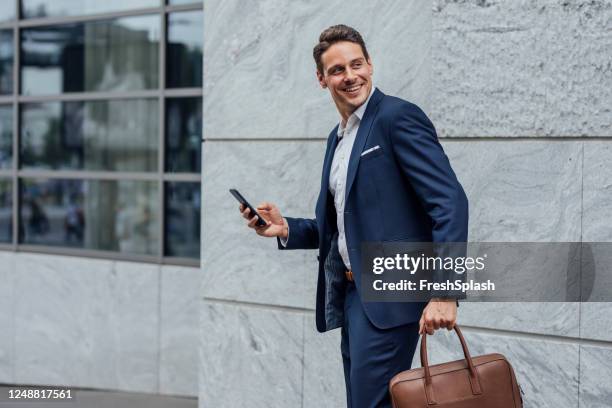 homme d’affaires beau de sourire dans un costume bleu vérifiant des médias sociaux sur son smartphone sur la rue - regarder par dessus son épaule photos et images de collection