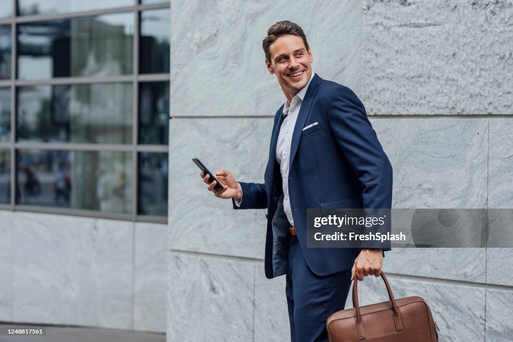 Smiling Handsome Businessman in a Blue Suit Checking Social Media on his Smartphone on the Street