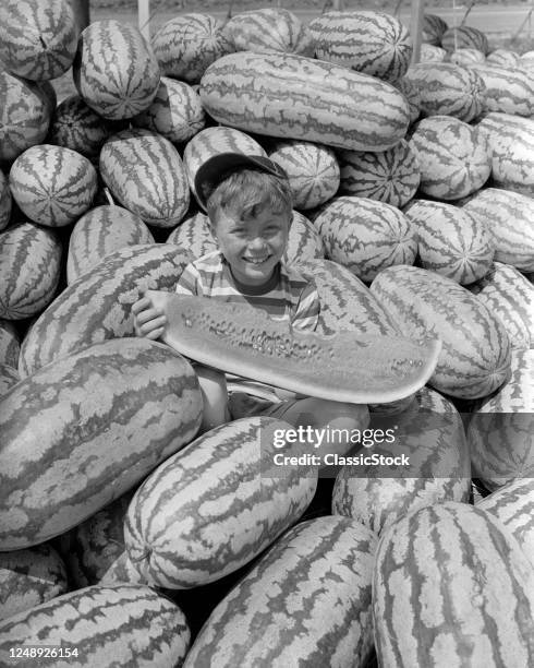 1940s 1950s Smiling Boy Wearing Ball Cap Looking At Camera Sitting In Pile Of Watermelons Eating A Large Slice Of One