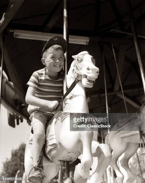 Eager Smiling Boy Wearing Ball Cap And Striped T-Shirt Riding On Carved Wooden Carousel Horse