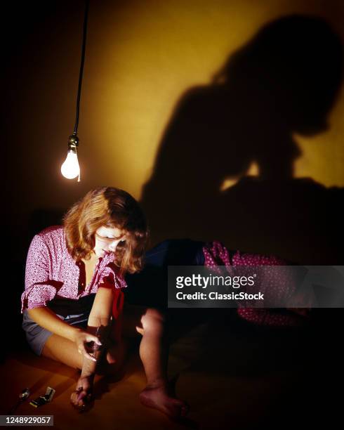 1960s 1970s Staged studio set-up of woman sitting Under Bare Bulb pretending to inject drugs illustrating drug use heroin a male companion is...