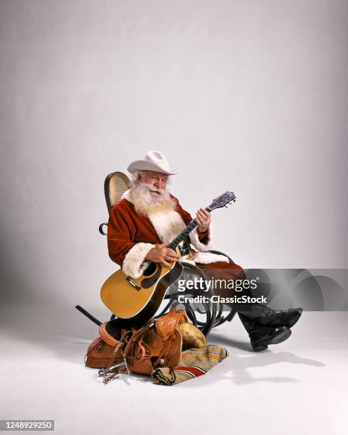 1980s Cowboy Santa Claus Looking At Camera Sitting In Bentwood Rocker Beside Western Saddle Playing Guitar Wearing White Hat