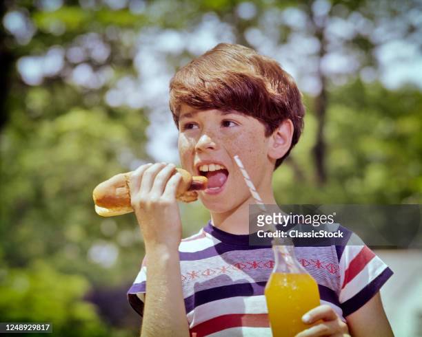 1970s 1980s Brunette Boy Freckle-Faced Wearing A Striped T-Shirt Biting Into Hot Dog Holding An Orange Soda With Paper Straw