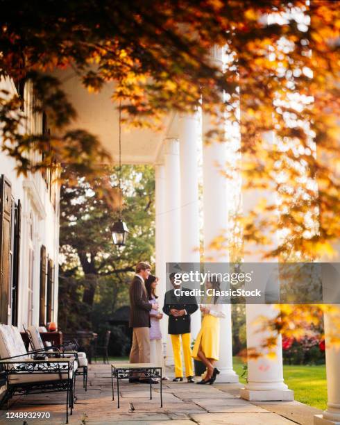 1970s Two Well-To-Do Couples Enjoying Having Cocktails Standing Talking On Patio Of Upscale House With Columns Autumn Sunlight