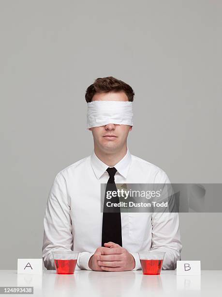 studio portrait of young man with blindfold sitting in front of two glasses with red liquid - taste test stock pictures, royalty-free photos & images