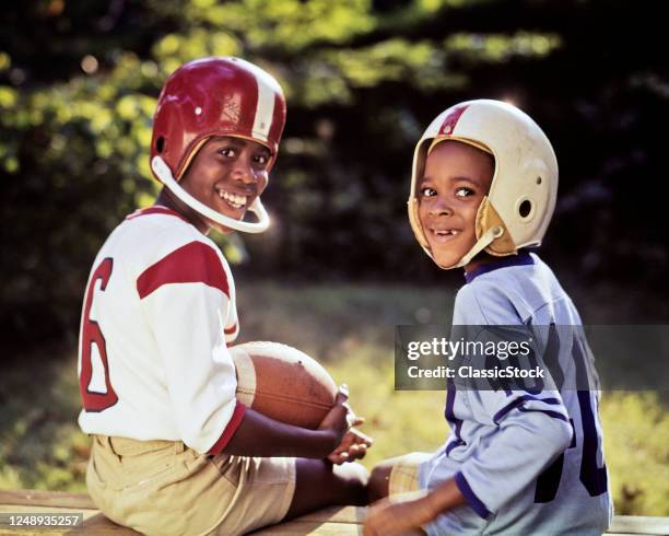 1970s Two Smiling African American Boys Smiling Looking At Camera Over Shoulders Wearing Football Jerseys Helmets Holding Ball