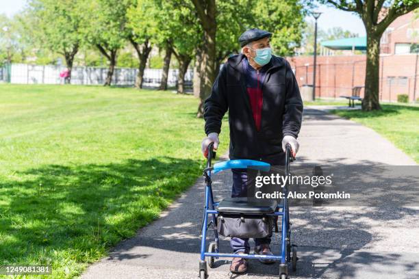 portrait of the senior 90-years-old man wearing protective mask and gloves because of the covid-19 pandemic walking outdoor in a park using a walker and maintaining social distancing. - alex potemkin coronavirus stock pictures, royalty-free photos & images