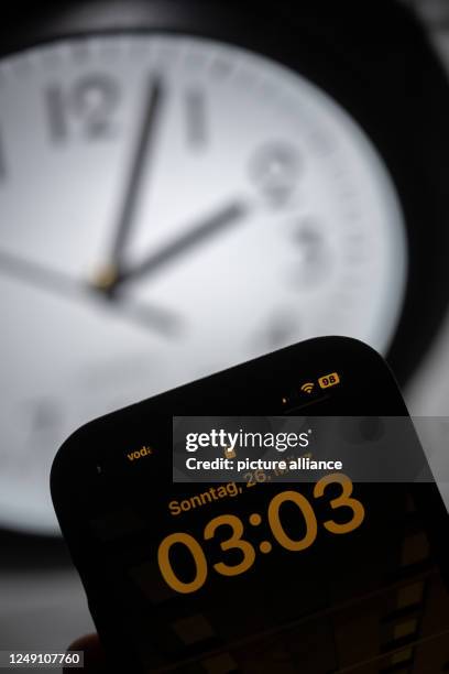 March 2023, Hesse, Gießen: A smartphone displays the time "03:03", while on an analog clock it is still 02:03. On March 26, the clocks will be set...