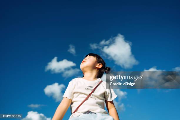 cute little asian girl with eyes closed enjoying the warmth of sun and the gentle breeze against beautiful blue sky on a lovely sunny day - child thinking stock-fotos und bilder