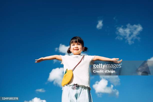 happy little asian girl smiling joyfully holding her hands up and imagines flying in the air against beautiful blue sky on a lovely sunny day - only kids at sky stockfoto's en -beelden