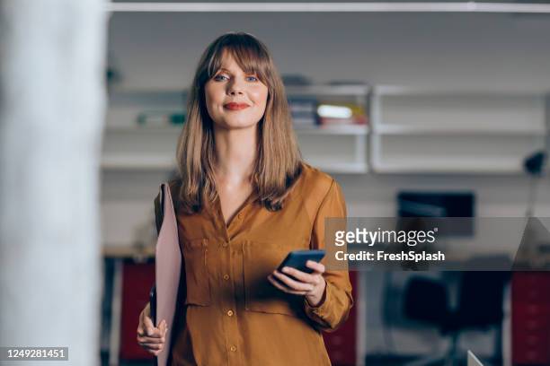 portrait of a beautiful blonde businesswoman walking through the office and using her smartphone - standing with laptop imagens e fotografias de stock