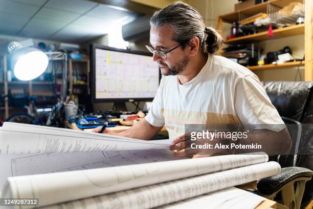 the mature, 50-years-old caucasian long-haired man working with architectural drawings in the home office located in a basement. - 50 54 years imagens e fotografias de stock