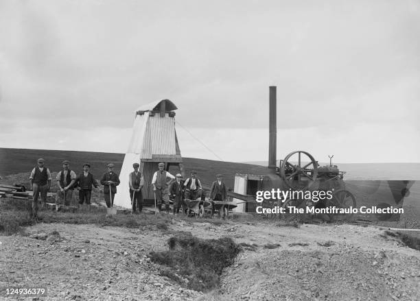 Group of young and old workmen with various tools, shovels and wheelbarrows standing close to a corrugated workshed and large industrial steam...