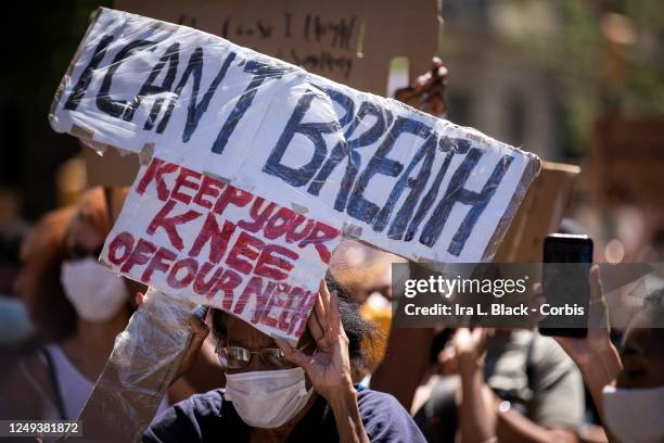 An elderly protester wearing a mask peeks through their homemade sign on a box that is held up and says, "I Can't Breath Keep Your Knee Off Our Neck"...