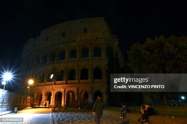 Pedestrians walk next to the ancient Colosseum, with the lights of the monument switched off as part of the Earth Hour initiative, in Rome on March...