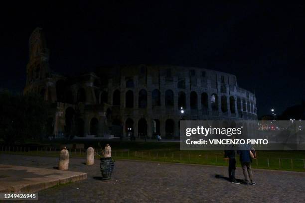 Pedestrians walk next to the ancient Colosseum, with the lights of the monument switched off as part of the Earth Hour initiative, in Rome on March...