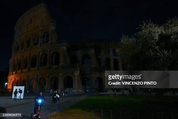 Pedestrians walk next to the ancient Colosseum, with the lights of the monument switched off as part of the Earth Hour initiative, in Rome on March...