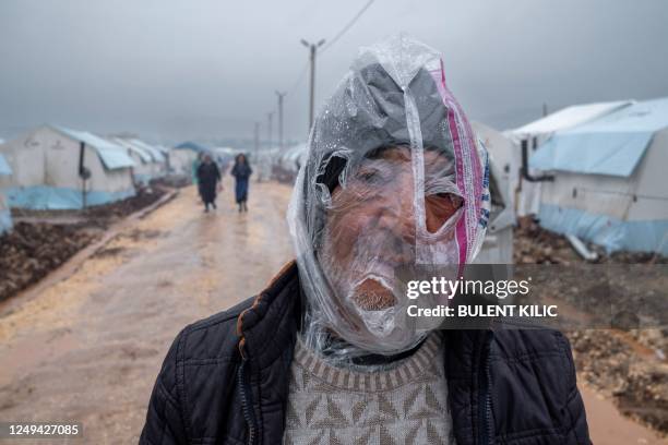 Man wears a plastic bag over his head to protect himself from the rain as he walks along a muddy road past tents set up to home displaced people...