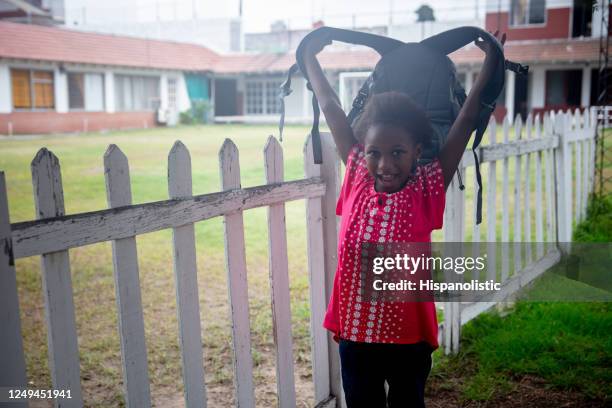 cute little black girl holding her backpack over her head while smiling at camera standing next to the school's fence - argentina girls stock pictures, royalty-free photos & images