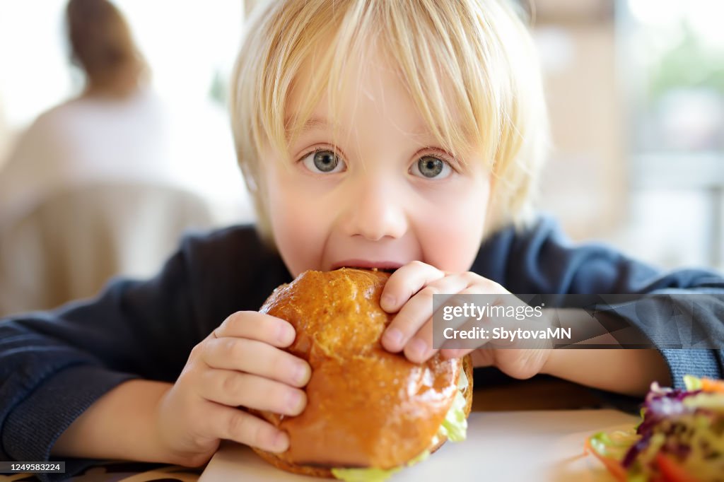 Cute blonde boy eating large hamburger at fastfood restaurant. Unhealthy meal for kids. Junk food.