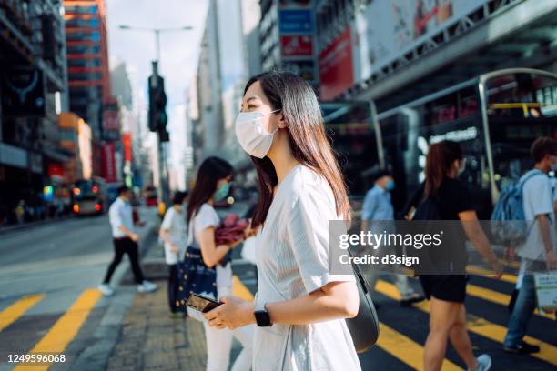 young asian woman with protective face mask using smartphone while commuting in downtown city street against crowd of pedestrians and city buildings - china coronavirus 個照片及圖片檔