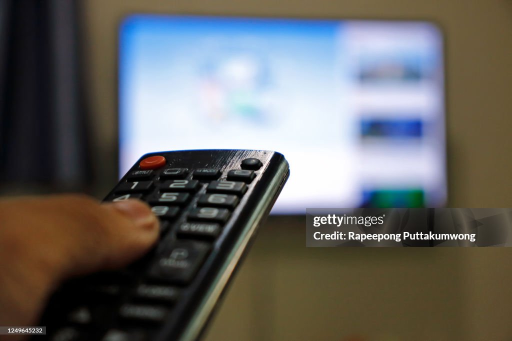 Hand holding remote control of television. Man watching TV at home and using remote control.