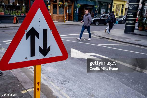 Two-way street traffic straight ahead sign interacts with passing people and an arrow painted on the road on 27th March 2023 in London, United...