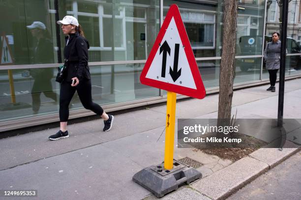 Two-way street traffic straight ahead sign interacts with passing people and an arrow painted on the road on 27th March 2023 in London, United...