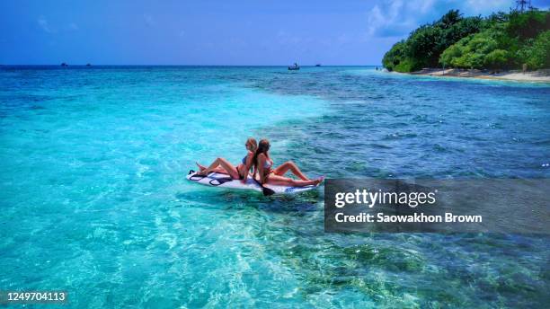 two female models sunbathing on top of a paddle board in an exotic beach with transparent waters - asian swimsuit models stock pictures, royalty-free photos & images