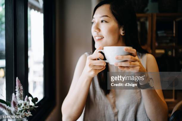 beautiful smiling young asian woman sitting in a cafe enjoying a cup of coffee and looking away through window - breakfast meeting stock-fotos und bilder