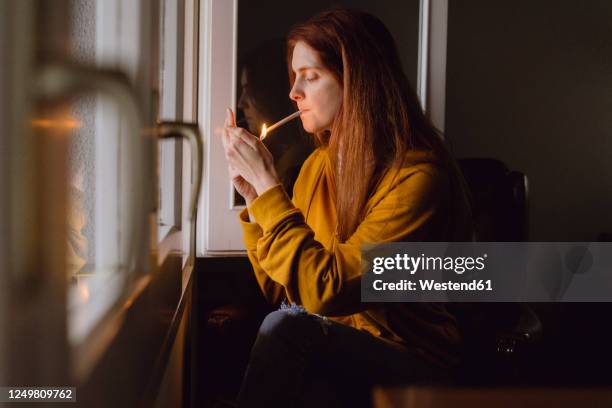 redheaded woman sitting at open window lighting joint - joint body part stock-fotos und bilder