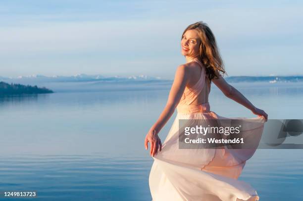 rear view portrait of smiling beautiful woman in dress standing at lakeshore on sunny day - women in see through dresses stock-fotos und bilder