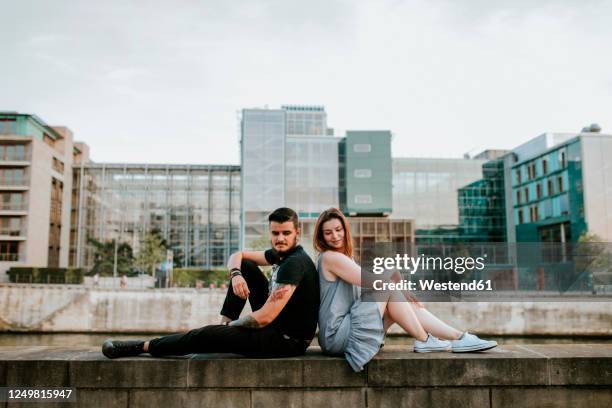 young couple sitting on a wall at the riverbank, berlin, germany - back to back stockfoto's en -beelden